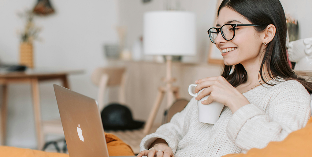Woman smiling while drinking coffee and using a laptop in a clean living room, representing the benefits of hiring cleaning services in Boca Raton.