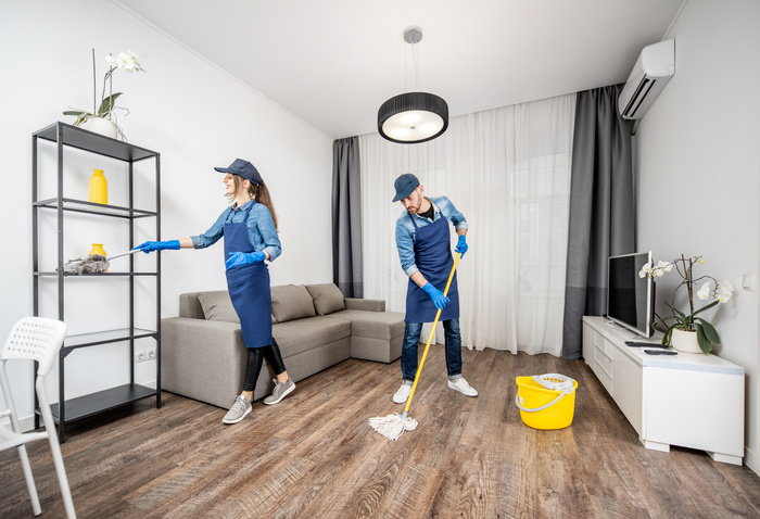 Professional cleaners in blue uniform washing floor and wiping dust from the furniture in the living room of the apartment. Cleaning service concept